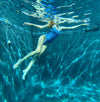 Photo is taken underwater of a girl in a bathing suit. Model wears scoop necked, spaghetti strap one piece. One piece has a baby blue, dark blue and light pink stripe print. The back features a low cut scoop that accentuates the back. Bathing suit is made sustainably out of recycled fishnets