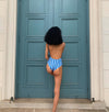 Model wears scoop necked, spaghetti strap one piece. One piece has a baby blue, dark blue and light pink stripe print. The back features a low cut scoop that accentuates the back. Bathing suit is made sustainably out of recycled fishnets