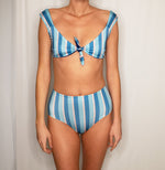 Model wears light blue, dark blue and light pink striped bikini. The bikini bottom is high waisted and full coverage. Each suit is made sustainable out of recycled fishnets